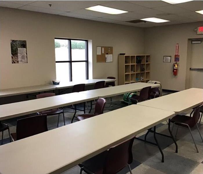 Cafeteria with 3 long tan tables and maroon chairs scooted in. Bulletin board and fire extinguisher on wall. 